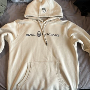 Beige hoodie från Sail Racing S - Snygg beige hoodie från Sail Racing i storlek S. Tröjan har svart logotyptryck på bröstet och huvan, samt praktisk känguruficka framtill. Mjuk insida och justerbar snörning i huvan gör den extra skön. Perfekt för chill dagar och streetwear.