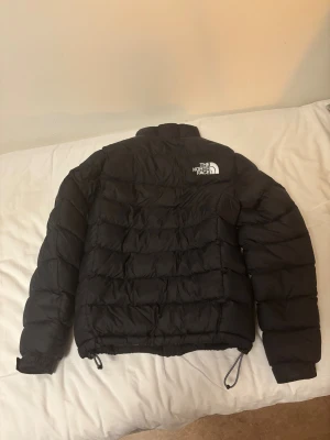 Svart pufferjacka från The North Face - Svart pufferjacka från The North Face med hög krage och synligt logotryck på bröstet. Jackan har dragkedja framtill, snörning i nederkant och två sidofickor. Den är quiltad och har långa ärmar, perfekt för kalla dagar.