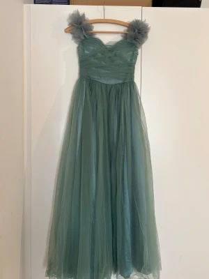 Grön långklänning med tyllaxlar - Drömmig grön långklänning med voluminösa tyllaxlar och omlottliv. Klänningen har ett svepande skirt tyg och snörning i ryggen för extra snygg passform. Perfekt för bal eller student! Materialet är lätt och ger en elegant känsla.
