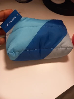 Blå randig necessär från Estée Lauder - Säljer en mjuk och quiltad necessär från Estée Lauder i olika blå nyanser med randigt mönster. Väskan har dragkedja upptill och är tillverkad i syntetmaterial. Perfekt för att förvara smink eller hudvårdsprodukter när du är på språng. Innehåller en 30ml Perfectly Clean Multi-Action Foam Cleanser/Purifying Mask, en 7ml DayWear kräm, en 5ml Advanced Night Repair kräm och en 7ml Advanced Night Repair serum.