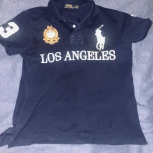 Mörkblå Polo Ralph Lauren Los Angeles piké - Snygg mörkblå pikétröja från Polo Ralph Lauren med vit Los Angeles-tryck, stort polospelare-logo och broderad emblem på bröstet. Nummer 3 i vitt på både ärm och rygg. Klassisk krage och korta ärmar, tillverkad i bomull. Perfekt för dig som gillar sportig och stilren look. Piken är en limited edition tröja som såldes under 2010-talet i mycket bra skick.