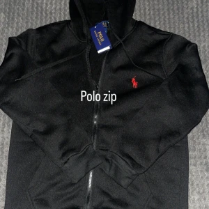 Svart hoodie från Polo Ralph Lauren - Svart hoodie från Polo Ralph Lauren med röd broderad logga på bröstet. Tröjan har huva med snörning, hel dragkedja och ribbade muddar vid ärmslut och nederkant. Perfekt för en chill och stilren look.