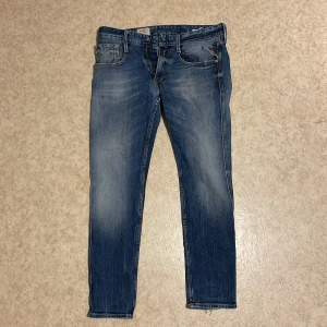 Replay Anbass blå jeans slim fit - Snygga Replay Anbass jeans i klassisk blå tvätt med slitningar och smal passform. Jeansen har fem fickor, Replay-logga på bakfickan och läderpatch i midjan. Perfekt för dig som gillar en modern och stilren look.