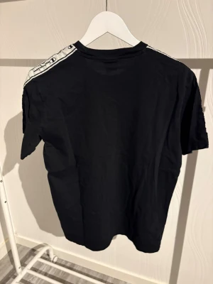 Svart Champion t-shirt med logoband - Svart t-shirt från Champion med vit logga på bröstet och coola logoband längs axlarna. Klassisk rund hals och korta ärmar. Materialet är mjuk bomull som känns skönt mot huden. Perfekt för dig som gillar sportig streetstyle och vill ha något som sticker ut.