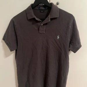 Mörkgrå polo t-shirt från Ralph Lauren - Säljer en mörkgrå polo t-shirt från Ralph Lauren med klassisk krage och två knappar framtill. T-shirten har korta ärmar och den ikoniska broderade loggan på bröstet. Tillverkad i mjuk bomull som är skön mot huden.