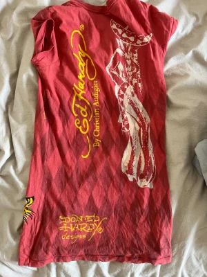 Röd Ed Hardy t-shirt med tryck - Säljer en röd t-shirt från Ed Hardy med coolt blommönster och tigertryck framtill samt en stor cowboy-figur på ryggen. T-shirten har korta ärmar, diamantmönster i bakgrunden och gula logotyper. Materialet är mjuk bomull och passformen är normal.