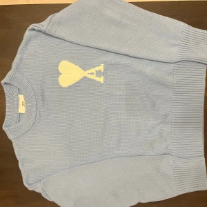 Ljusblå stickad tröja från Ami Paris - Säljer en ljusblå stickad tröja från Ami Paris med ikoniskt vitt hjärta och A-logga på bröstet. Tröjan har rund halsringning, ribbade muddar och är långärmad. Perfekt för dig som vill ha en clean och stilren look med en touch av designer vibes.