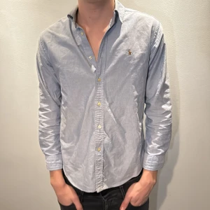 Blå skjorta från Ralph Lauren - Snygg ljusblå skjorta från Ralph Lauren med klassisk krage och broderad logga på bröstet. Skjortan har långa ärmar, knappar framtill och är tillverkad i mjuk bomull. Perfekt för dig som gillar stilrena och tidlösa plagg. Modellen är 186cm och väger 70 kg 