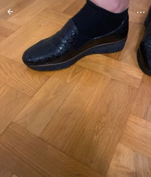 Svarta loafers från Rieker med mönster - Säljer ett par svarta loafers från Rieker med glansig yta och präglat mönster som påminner om ormskinn. Skorna har en chunky platt sula och är tillverkade i syntetmaterial. Perfekta för dig som vill ha både stil och komfort.