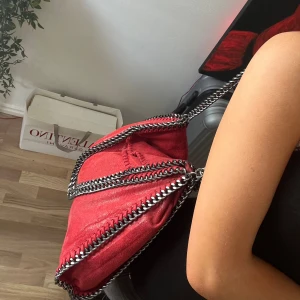 Röd väska med kedjedetaljer - Cool röd axelväska från Stella McCartney med glansig yta och kraftiga silverfärgade kedjor längs kanterna och som axelrem. Väskan har en mjuk, slouchy form och är tillverkad i syntetmaterial som liknar mocka. Perfekt för dig som vill sticka ut med en statementväska.