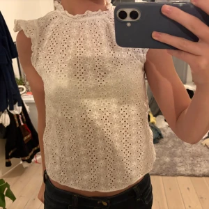 Zara blus med volang - Säljer en söt vit blus med broderade blommor och volangdetaljer vid axlar och hals. Blusen är ärmlös och har en lätt croppad passform. Materialet känns som bomull och är luftigt.