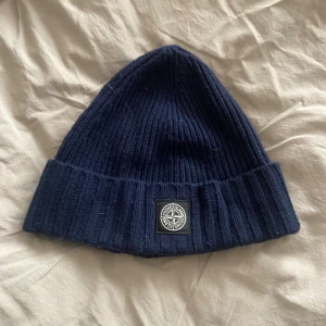 Stone Island  ribbad mössa junior - Snygg marinblå ribbstickad mössa från Stone Island med klassisk logopatch framtill. Mössan har bred uppvikt kant och är tillverkad i mjukt stickat material som håller dig varm och stilren under kalla dagar.