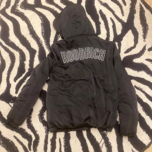 Svart pufferjacka från Hoodrich - Svart pufferjacka från Hoodrich med stor logga på ryggen och mindre logga framtill. Jackan har huva, dragkedja och en loose passform. Perfekt för kalla dagar och har en stilren streetkänsla som sticker ut.