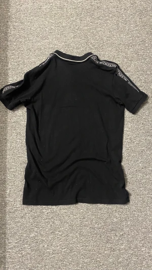 McKenzie Polo Shirt - Svart pikétröja från McKenzie med vit piping på kragen och tryckt logga på bröstet. Axlarna har ett band med McKenzie-tryck för extra streetkänsla. Tröjan har korta ärmar och knappar framtill. Materialet är mjuk bomull som är skön mot huden.