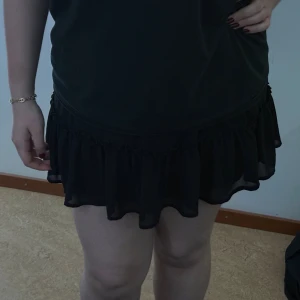 Svart volangkjol i chiffong - Säljer en svart kortkjol med volangkant i lätt och flödande chiffongmaterial. Kjolen har en enkel design med elastisk midja och volanger som ger en snygg och lekfull look. Perfekt för dig som gillar att sticka ut med en feminin stil.