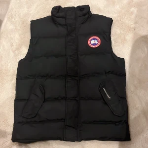 Svart dunväst från Canada Goose - Svart dunväst från Canada Goose med klassisk hög krage och broderad logotyp på bröstet. Västen har två fickor med lock och diskret Canada Goose-märke på ena fickan. Perfekt för kalla dagar och har en stilren, quiltad design.