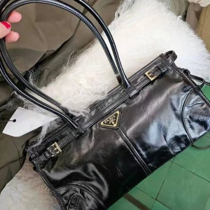 Paris axelväska i skinn med spännen - Snygg axelväska från Paris med stilrena dubbla handtag och guldfärgade spänndetaljer. Finns i svart, rosa och grått skinn med en elegant, avlång form och triangulär logga framtill. Perfekt för dig som vill ha en trendig och rymlig väska med lyxig känsla.