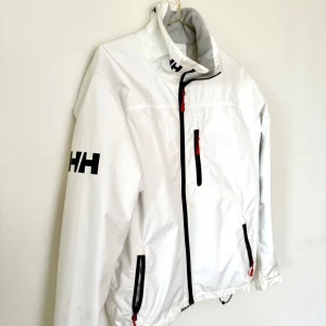 Vit vindjacka från Helly Hansen S - Vit vindjacka från Helly Hansen med svart HH-logga på ärmen och detaljer i svart och rött vid dragkedjorna. Jackan har hög krage, tre fickor med dragkedja och meshfoder på insidan. Perfekt för dig som gillar sportig och clean stil. Passar medium