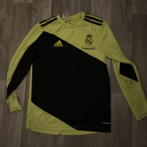 Real Madrid långärmad målvaktströja - Säljer en långärmad Real Madrid målvaktströja från Adidas i neongul och svart. Tröjan har svarta axelränder, vadderade partier på armbågarna och tryckt klubbmärke samt Adidas-logga på bröstet. Materialet är lätt och andas, perfekt för fotbollsträning. Fick denna när jag var och provtränade för dem