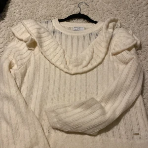 Vit stickad tröja med volang Holly & Whyte - Supersöt vit stickad tröja från Holly & Whyte med ribbat mönster och bred volang längs axlar och halsringning. Långärmad och mjuk, perfekt för dig som gillar romantiska detaljer och vill ha en mysig look. Passar dig som vill ha något extra till garderoben.