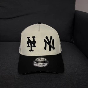 New Era vit och svart keps NY Mets - Snygg keps från New Era i vit och svart med broderade NY Mets och Yankees-loggor framtill. Justerbar snapback baktill och 25th Anniversary World Series patch på sidan. Klassisk 9FORTY-modell i bomull, perfekt för dig som gillar baseball och streetstyle.