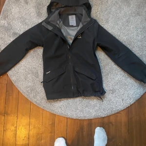 Svart vindjacka från Haglöfs - Svart vindjacka från Haglöfs i juniorstorlek 164. Jackan har huva, dragkedja framtill och två stora fickor med lock. Perfekt för blåsiga dagar och outdoor-aktiviteter. Snygg och funktionell design med diskret logga på bröstet. Pris kan diskuteras! Skriv vid frågor! Denna passar mig perfekt då jag är 175 cm och väger 55 kg. Den går att ställa in nere i midjan