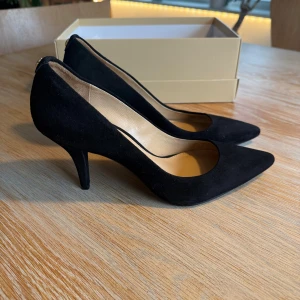 Svarta pumps från Michael Kors - Snygga svarta pumps från Michael Kors. Skorna har spetsig tå, låg klack och är tillverkade i mjuk mocka. På baksidan finns en liten guldfärgad detalj med logga. Perfekt för dig som gillar stilrena och eleganta skor. Storlek 38 men små i storleken, jag skulle säga stl 37. Nypris 1600:- 