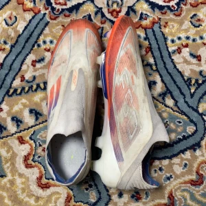 Adidas F50 fotbollsskor i vitt och orange - Säljer ett par Adidas F50 fotbollsskor med en snygg design i vitt, orange och blått. Skorna har platt sula med dobbar för gräsplan och är tillverkade i syntetmaterial. De har en strömlinjeformad siluett utan snörning för en tight passform och maxad speed på planen. Använda av ett allsvenskt proffs i Degerfors IF