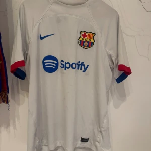  FC Barcelona fotbollströja 2023/24 - Säljer en vit FC Barcelona fotbollströja från Nike i storlek L (säsong 2023/24) Tröjan har korta ärmar med röda och blå detaljer, Barcelonas klubbmärke på bröstet och blå Nike-logga samt Spotify-tryck. Tillverkad i lätt och ventilerande polyester. Tröjan är lite missfärgad i tvätten därav några enstaka fläckar. Tröjan är köpt på Nike hemsidan, så denna är äkta (kolla bild). 
