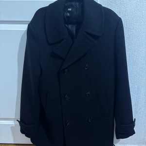 Svart skepparkavaj från H&M - Svart skepparkavaj från H&M med klassisk dubbelknäppning och bred krage. Jackan har två sidofickor och snygga detaljer vid ärmsluten. Materialet är mjukt och känns som ullblandning, perfekt för kyliga dagar. Stilren och tidlös design som funkar till många outfits.