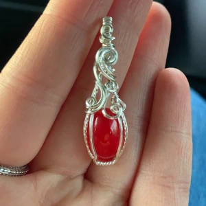 Halsbandshänge med röd sten och silvertråd - Unikt halsbandshänge med en intensivt röd, oval sten infattad i detaljerad silverfärgad tråd. Hängsmycket har snygga snirkliga mönster och en elegant, handgjord känsla. Perfekt för dig som vill sticka ut med något personligt och färgstarkt.