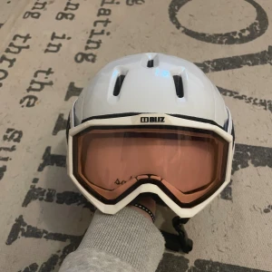 Vit Salomon skid/sporthjälm med Bliz goggles - Säljer en vit Salomon hjälm i storlek S med ventiler och mjuk, beige fuskpäls på insidan för extra komfort. Medföljer Bliz goggles med rosa tonade linser och vit ram. Perfekt för dig som vill ha både stil och skydd i backen.
