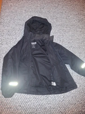 Svart vindjacka från Texstar, strl 152 - Svart vindjacka från Texstar med huva och reflexdetaljer på ärmar och rygg. Jackan har dragkedja framtill och är tillverkad i slitstark polyester. Perfekt för blåsiga dagar och har en clean, sportig look som funkar till de flesta outfits. Denna jacka är också starkt vattentät och perfekt för sport och andra aktiviteter.