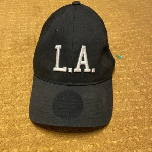 Svart keps med L.A. - Svart keps med böjd skärm och justerbar rem baktill. Framtill sitter en stor broderad L.A.-text i vitt som ger en urban vibe. Kepsen är tillverkad i bomull och har klassisk sexpanelsdesign med ventilationshål.