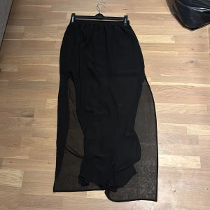 Svart långkjol med transparent nederdel - Svart långkjol med elastisk midja och snygg transparent nederdel. Kjolen har en slät, enkel design med ett lager som är mer täckande upptill och ett tunt, genomskinligt tyg nedtill. Perfekt för dig som gillar stilrena och moderna plagg. Splittad ca vid knähöjd på båda sidorna. OBS!! Tvättas innan!!
