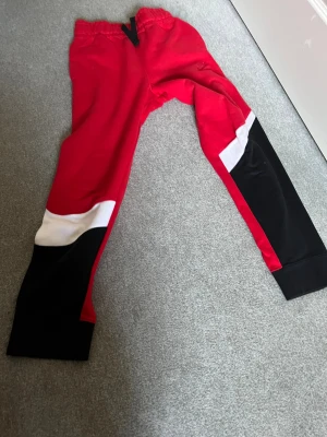 Nike röda mjukisbyxor med färgblock - Snygga röda mjukisbyxor från Nike med coola färgblock i svart och vitt på benen. Byxorna har elastisk midja med snörning och är gjorda i mjukt bomullsmaterial. Perfekta för chill eller träning, med en loose passform och sportig vibe.