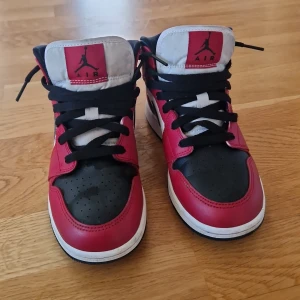Nike Air Jordan 1 Mid röd/svart/vit 36.5 - Säljer ett par Nike Air Jordan 1 Mid i klassisk röd, svart och vit färg. Skorna har svart snörning, perforerad tå och ikonisk Jumpman-logga på plösen. Tillverkade i skinn och syntet med vit sula och svart swoosh på sidan. Perfekta för dig som gillar streetstyle.