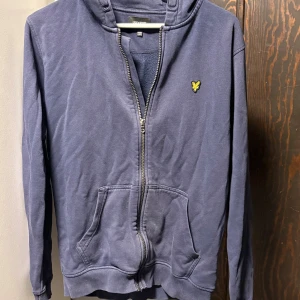 Marinblå hoodie från Lyle & Scott - Snygg marinblå hoodie från Lyle & Scott Junior med gul logga på bröstet. Tröjan har dragkedja, huva och två fickor framtill. Tillverkad i mjuk bomull och har långa ärmar. 