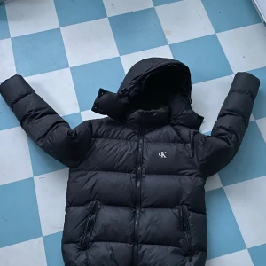 Svart pufferjacka från Calvin Klein - Svart pufferjacka från Calvin Klein med huva och dragkedja framtill. Jackan har två sidofickor med dragkedja och ett diskret CK-logo på bröstet. Den är quiltad och har en oversized look, perfekt för kalla dagar.