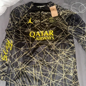 PSG långärmad fotbollströja Jordan M - Svart långärmad PSG fotbollströja från Jordan med gula detaljer och loggor. Tröjan har ett unikt grafiskt mönster med gula linjer över hela ytan, halv dragkedja framtill och Qatar Airways tryck. Tillverkad i lätt, tekniskt Dri-Fit material som andas.