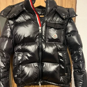 Svart Moncler pufferjacka  - Svart pufferjacka från Moncler med glansig finish, ikonisk logga på bröstet och praktiska fickor med dragkedja och knapp. Jackan har hög krage, lång ärm och snygga quiltade paneler. Perfekt för kalla dagar och har en modern, urban vibe. 