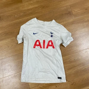 Tottenham Hotspur vit matchtröja Nike - Vit Tottenham Hotspur fotbollströja från Nike med klubbmärke och blå Nike-logga på bröstet samt röd AIA-sponsor. Klassisk V-ringning och korta ärmar. Tillverkad i lätt och ventilerande material, perfekt för träning eller match.