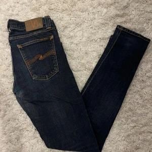 Mörkblå skinny jeans från Nudie Jeans - Snygga mörkblå fit jeans från Nudie Jeans med klassiska orangea sömmar och coola detaljer på bakfickorna. Modellen har smal passform och normal midja, tillverkade i stretchigt jeansmaterial för en bekväm känsla. Perfekt för dig som gillar stilrena och tajta jeans.
