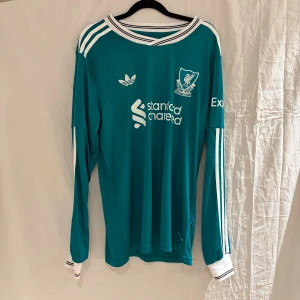 Liverpool långärmad fotbollströja Adidas - Liverpool långärmad fotbollströja från Adidas i turkosblått med vita detaljer, klubbmärke och sponsortryck framtill. Nummer 9 och namnet ISAK på ryggen. Klassiska Adidas-ränder på axlar och ärmar. Tillverkad i lätt och ventilerande polyester.