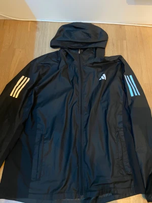 Svart vindjacka från Adidas med huva - Svart vindjacka från Adidas med huva och vita ränder på båda ärmarna. Perfekt för löpning med sportig look och RUNNING-detalj på insidan. Vill bli av med jackan, priset går såklart att diskutera. :) 
