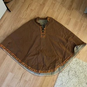 Brun poncho med broderad kant - Unik brun poncho med broderade detaljer i orange, svart och grönt längs kanten och vid halsen. Ponchon har snörning vid halsöppningen och är fodrad med grått tyg på insidan. Snörningen fungerar inte till 100%.