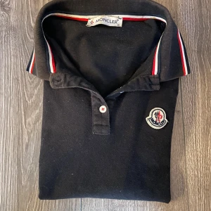 Svart Moncler pikétröja med röd/vit detalj - Svart pikétröja från Moncler med klassisk krage och knappar framtill. Tröjan har Moncler-logga på bröstet och snygga rödvita ränder på krage och ärmslut. Storlek S=155-165