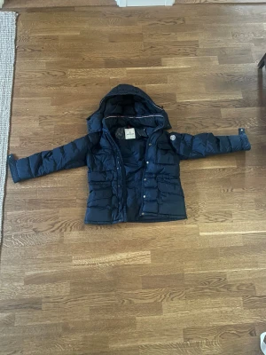 Mörkblå Moncler dunjacka med huva - Säljer en mörkblå dunjacka från Moncler med stor, avtagbar huva och flera fickor med knappar. Nypris: 12 000kr. Jackan har både dragkedja och knappar framtill, samt Moncler-logga på ärmen. Perfekt för kalla dagar och riktigt snygg till streetwear.