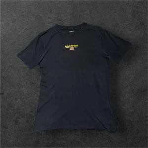 Svart Polo Sport t-shirt från Ralph Lauren - Snygg svart t-shirt från Polo Sport Ralph Lauren med klassisk logga och liten amerikansk flagga på bröstet. Modellen är kortärmad och har en rund hals. Tillverkad i mjuk bomull som känns skön mot huden. Perfekt för dig som gillar stilrena och sportiga plagg.