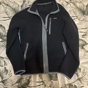 Mörkblå fleecejacka från Patagonia Junior XL - Mysig mörkblå fleecejacka från Patagonia i storlek Junior XL. Jackan har grå detaljer vid dragkedjor och krage, tre fickor med dragkedja och ett broderat Patagonia-märke på bröstet. Perfekt för kalla dagar och chill stunder. Junior XL=160-165CM skulle jag säga 